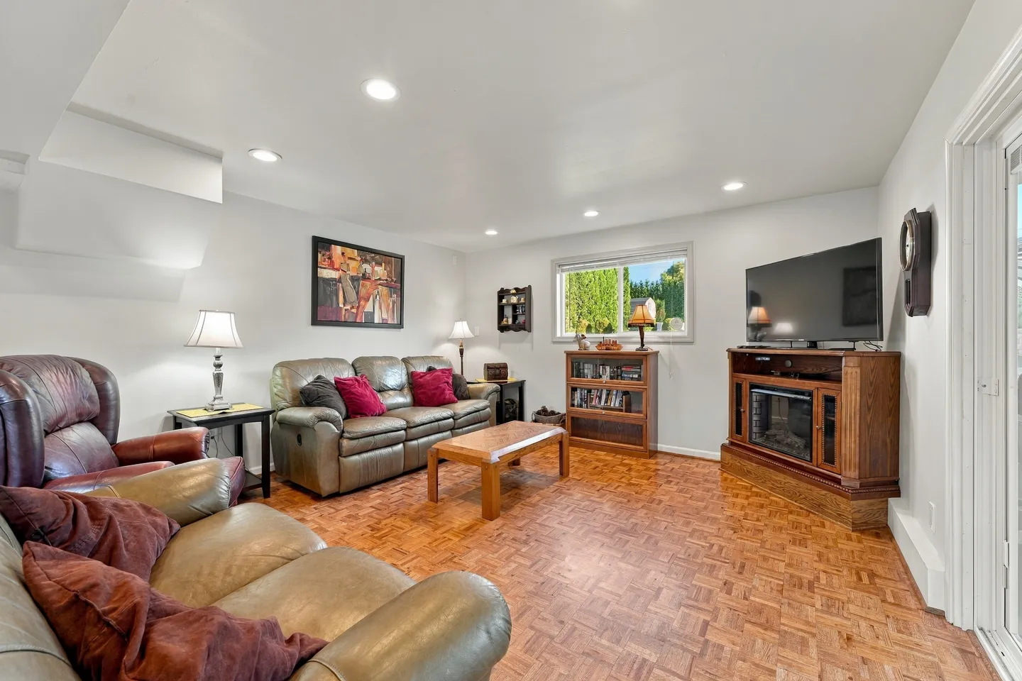 Living room with leather seating, parquet-style flooring, a corner television console with an electric fireplace, and a window.