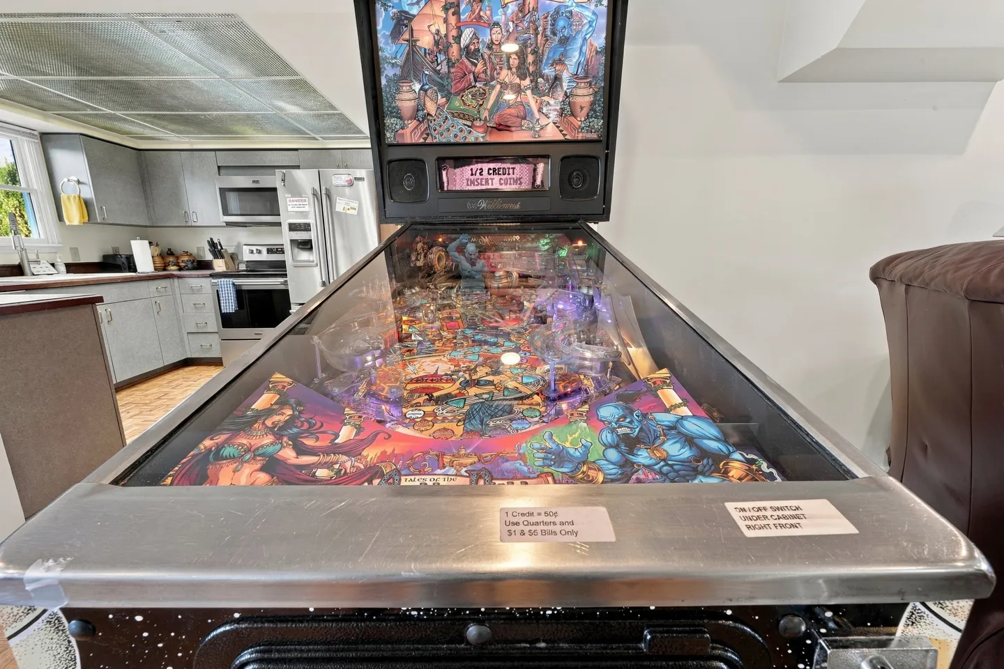 Close-up perspective of an illuminated pinball machine in an open-concept living space with a kitchen in the background.