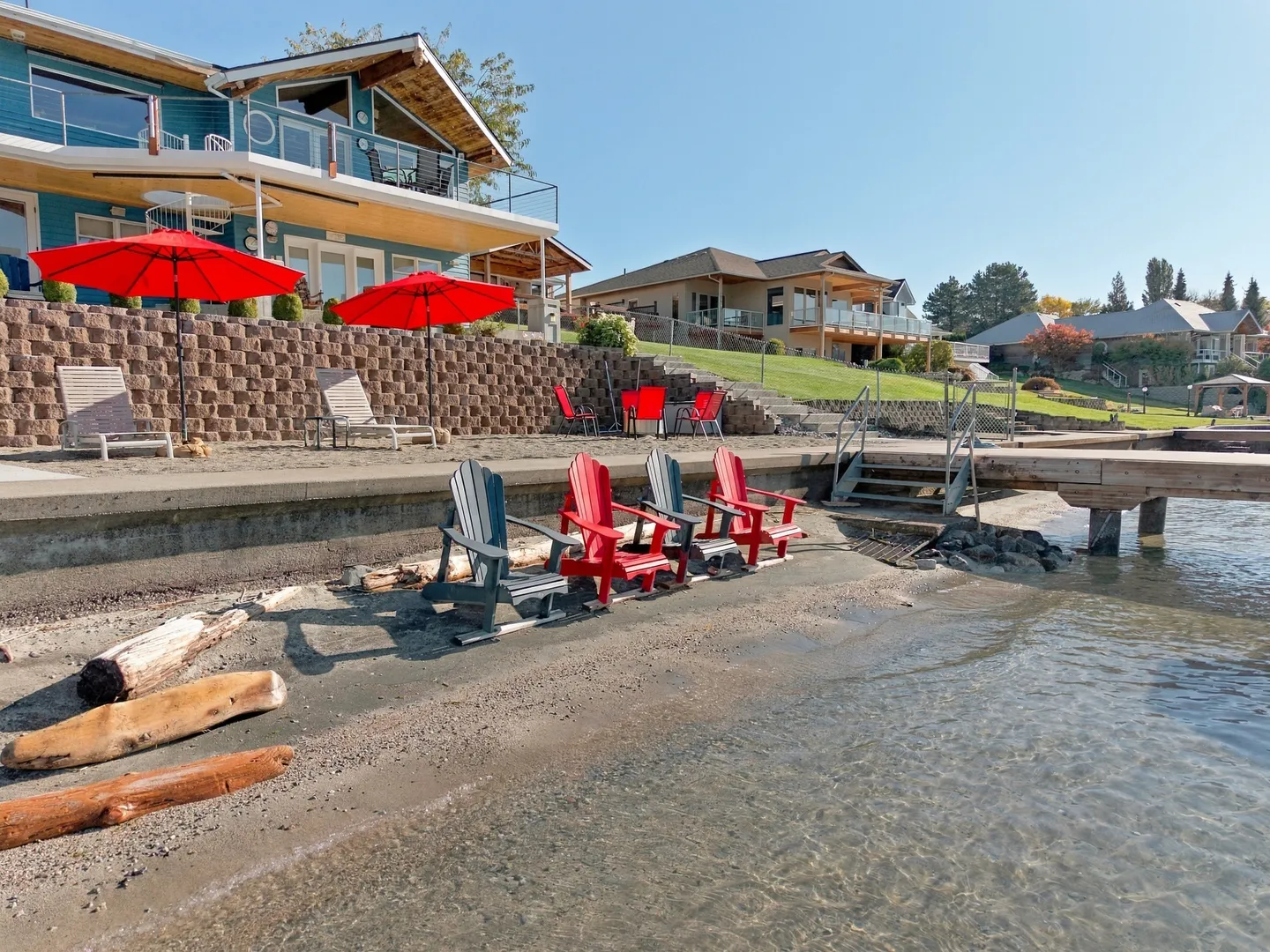 Lakeshore beach area in front of a multi-level house with Adirondack chairs on the sand and a dock extending into clear water.