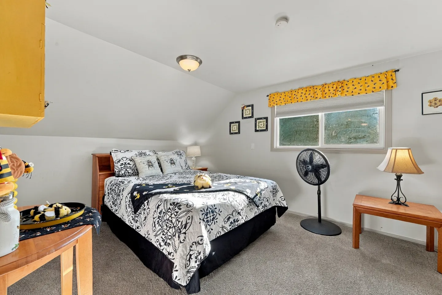 Bedroom with a patterned bedspread, sloped ceiling, window with a yellow valance, and carpeted floor.