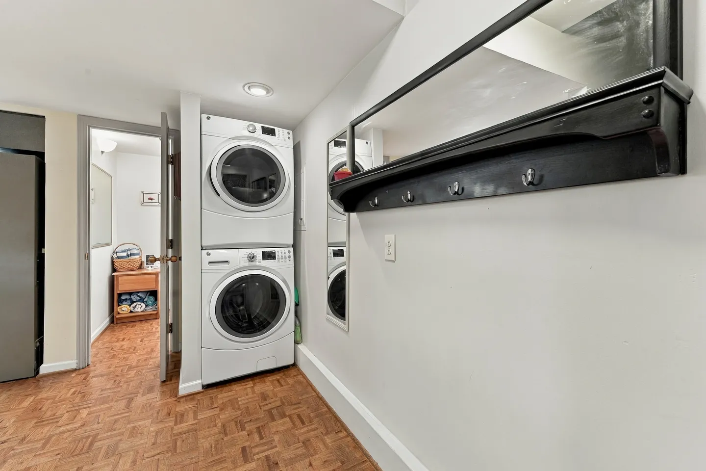 Hallway laundry area with stacked white washer and dryer, parquet wood flooring, and a wall-mounted coat rack with mirror.