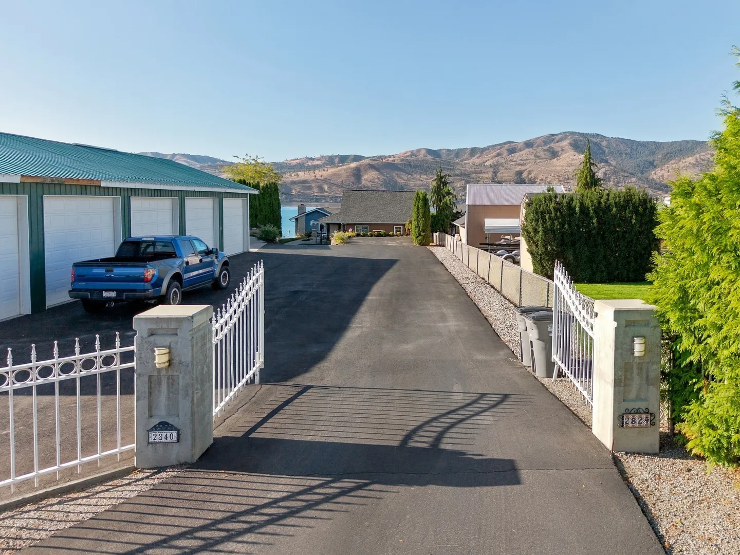 Open white iron gates at the entrance to a long paved driveway leading toward a house and distant water with hills in the background.