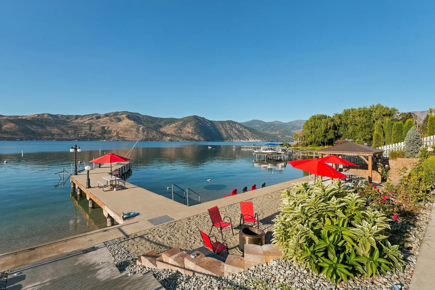 Lakeside patio and dock with red chairs, umbrellas, a fire pit, and mountain views across clear water.