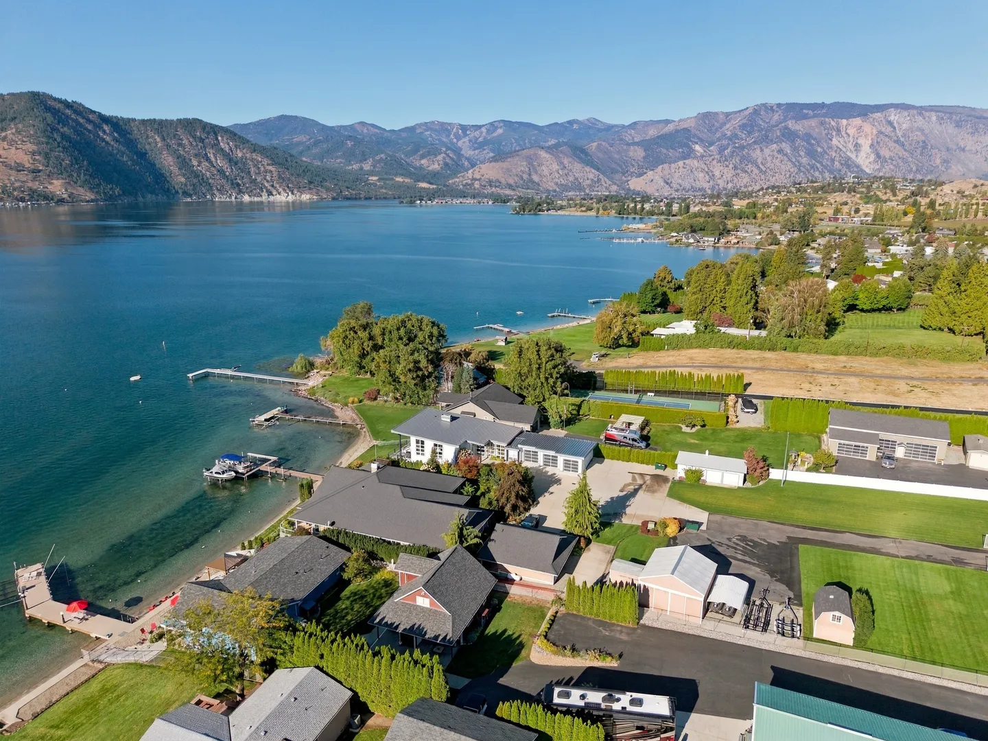 Aerial view of waterfront homes along a blue lake surrounded by mountains under a clear sky.