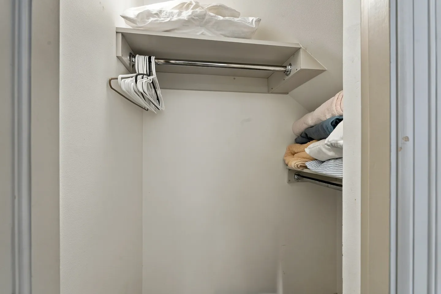 Interior view of a small closet with clothing racks, hangers, and folded linens on shelves.