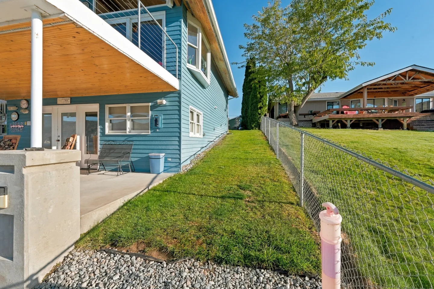 Exterior side view of a blue two-story house showing a sloping grassy lawn running alongside a chain-link fence, a ground-level concrete patio, and an upper deck.