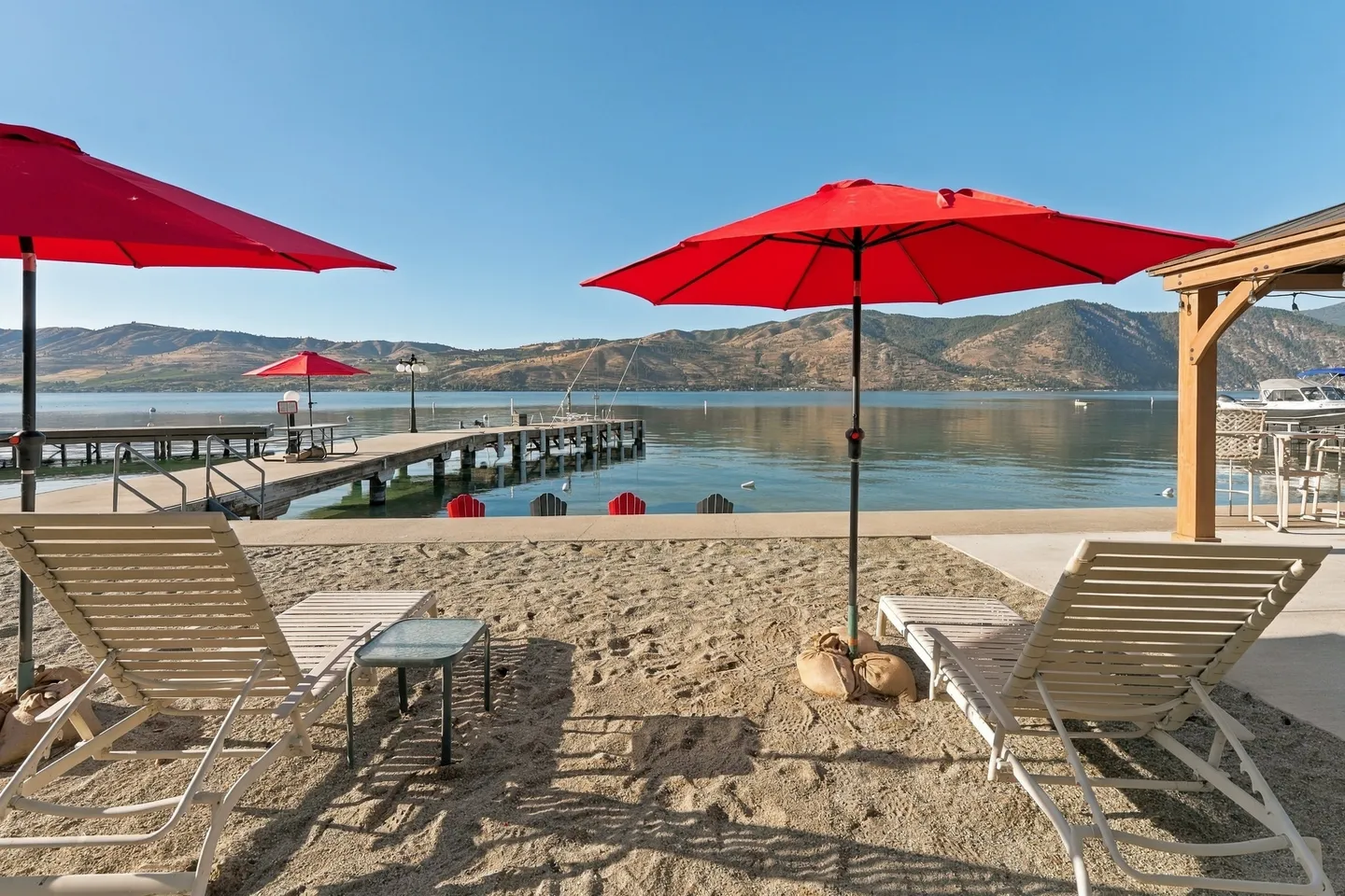 Lounge chairs and bright red umbrellas on a sandy beach overlooking a lake, wooden dock, and distant hills.