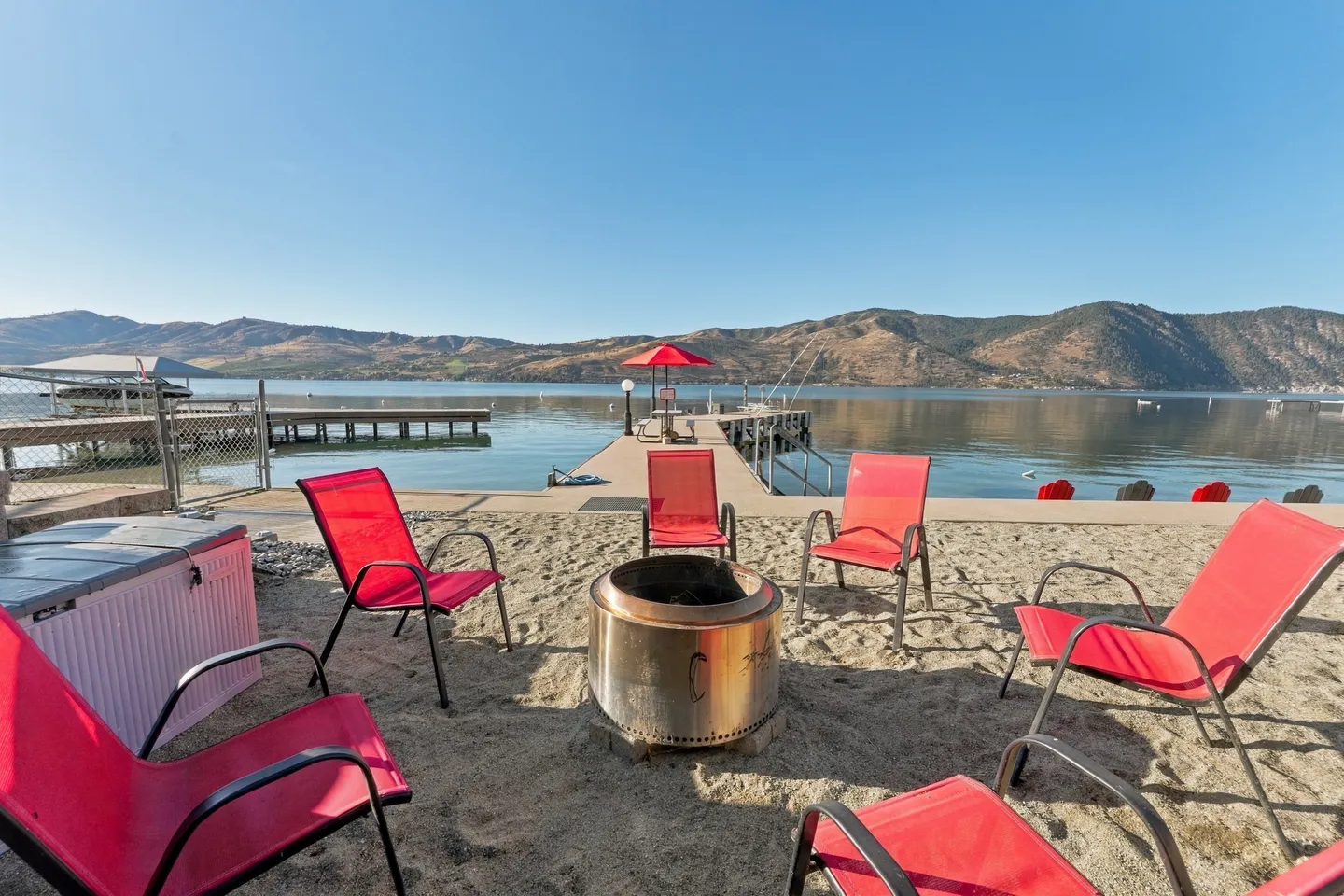 A sandy fire pit area with red chairs overlooking a lake dock and mountains under a clear blue sky.