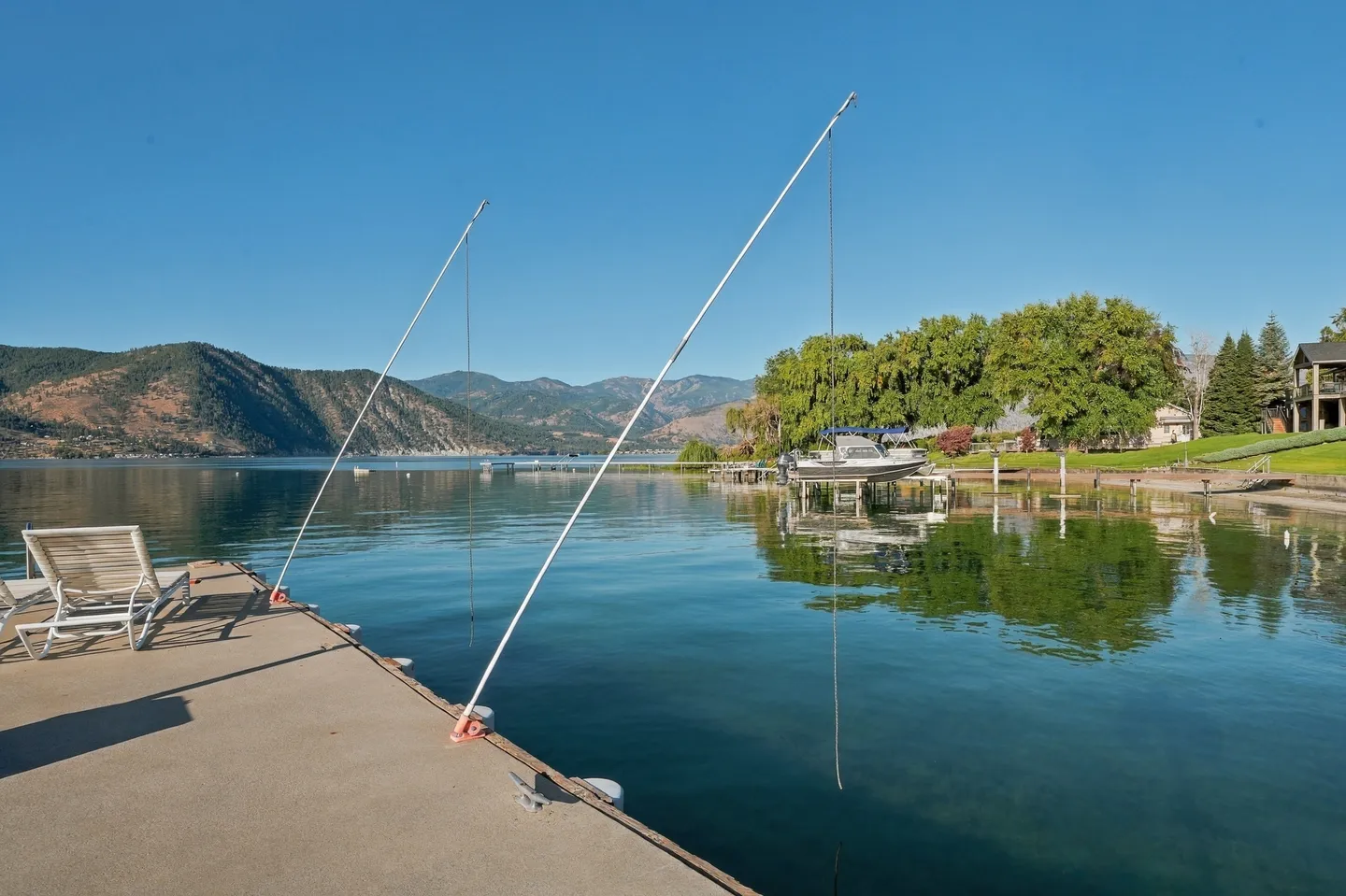 A floating dock extending into clear blue water with lounge chairs and mooring whips, looking toward a tree-lined shore and mountains under a clear sky.