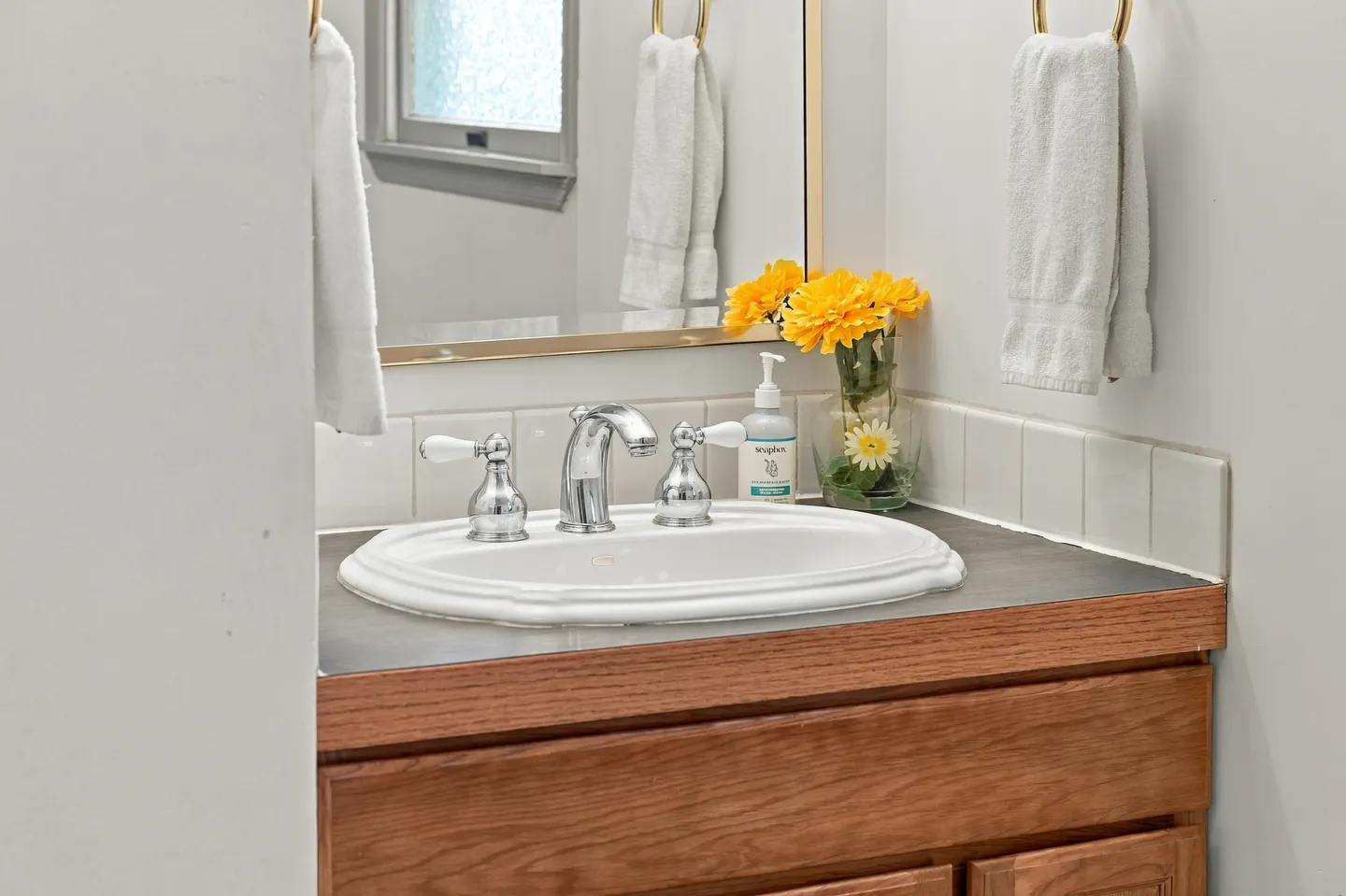 Bathroom vanity with a white drop-in sink, wooden cabinet, chrome and white faucet handles, and a vase of yellow flowers.