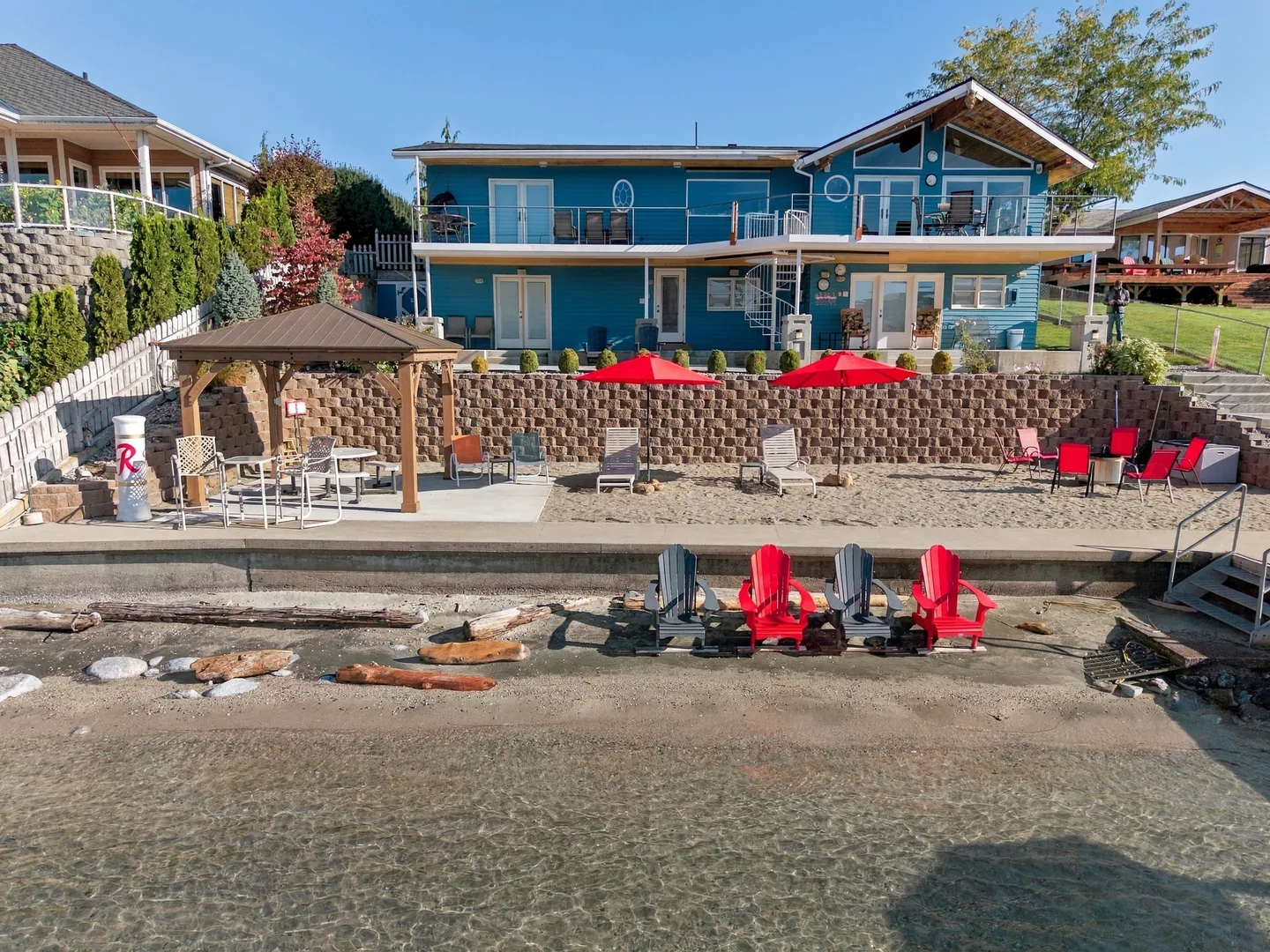 Rear exterior of a two-story blue waterfront house with a sandy beach, gazebo, and outdoor seating.