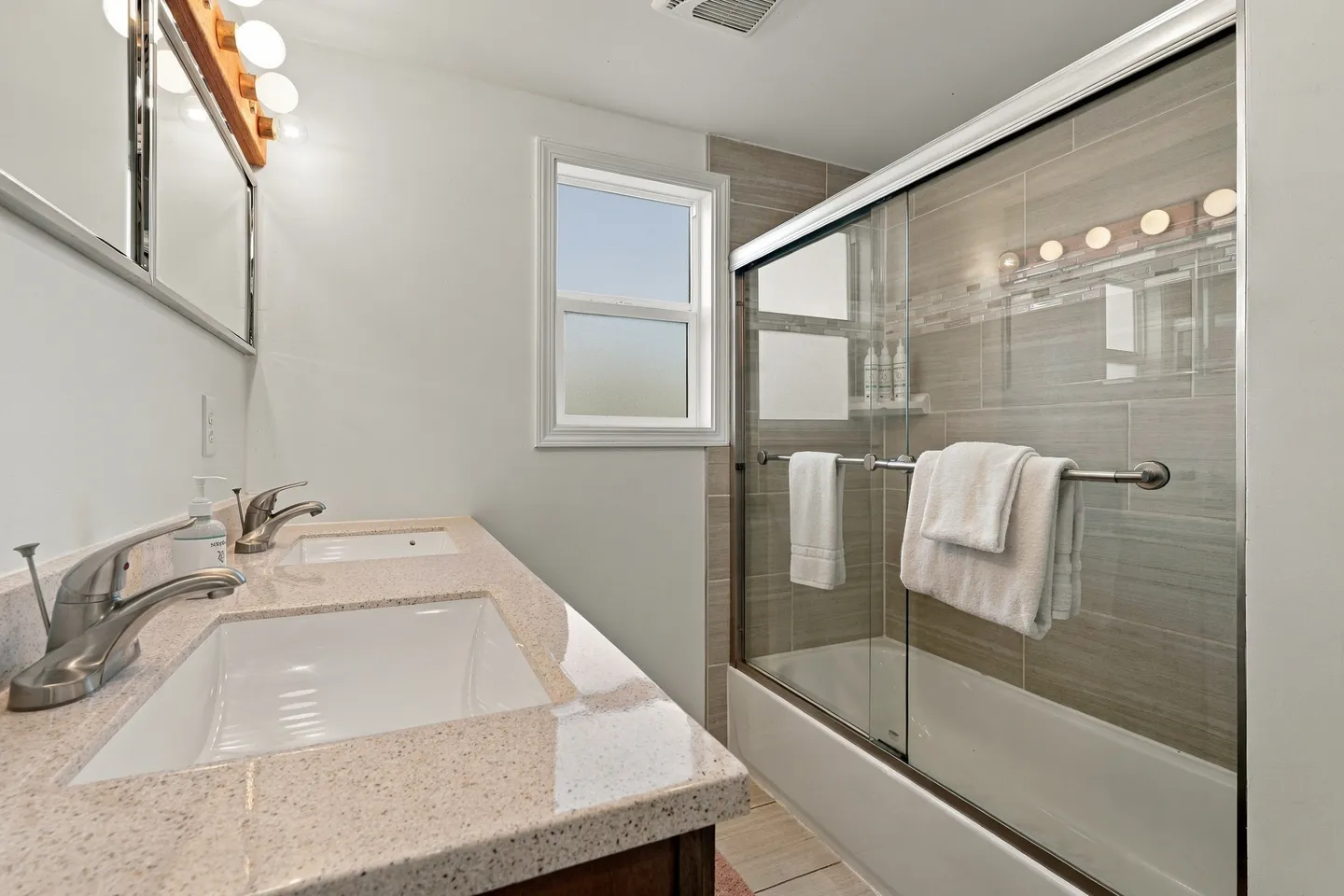 Bathroom showing a double-sink vanity in the foreground and a bathtub with sliding glass doors and tiled walls.