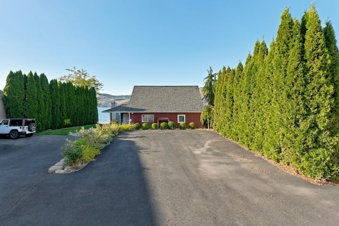 A wide paved driveway leading to a red house, lined on both sides by tall green arborvitae hedges with water visible in the background.