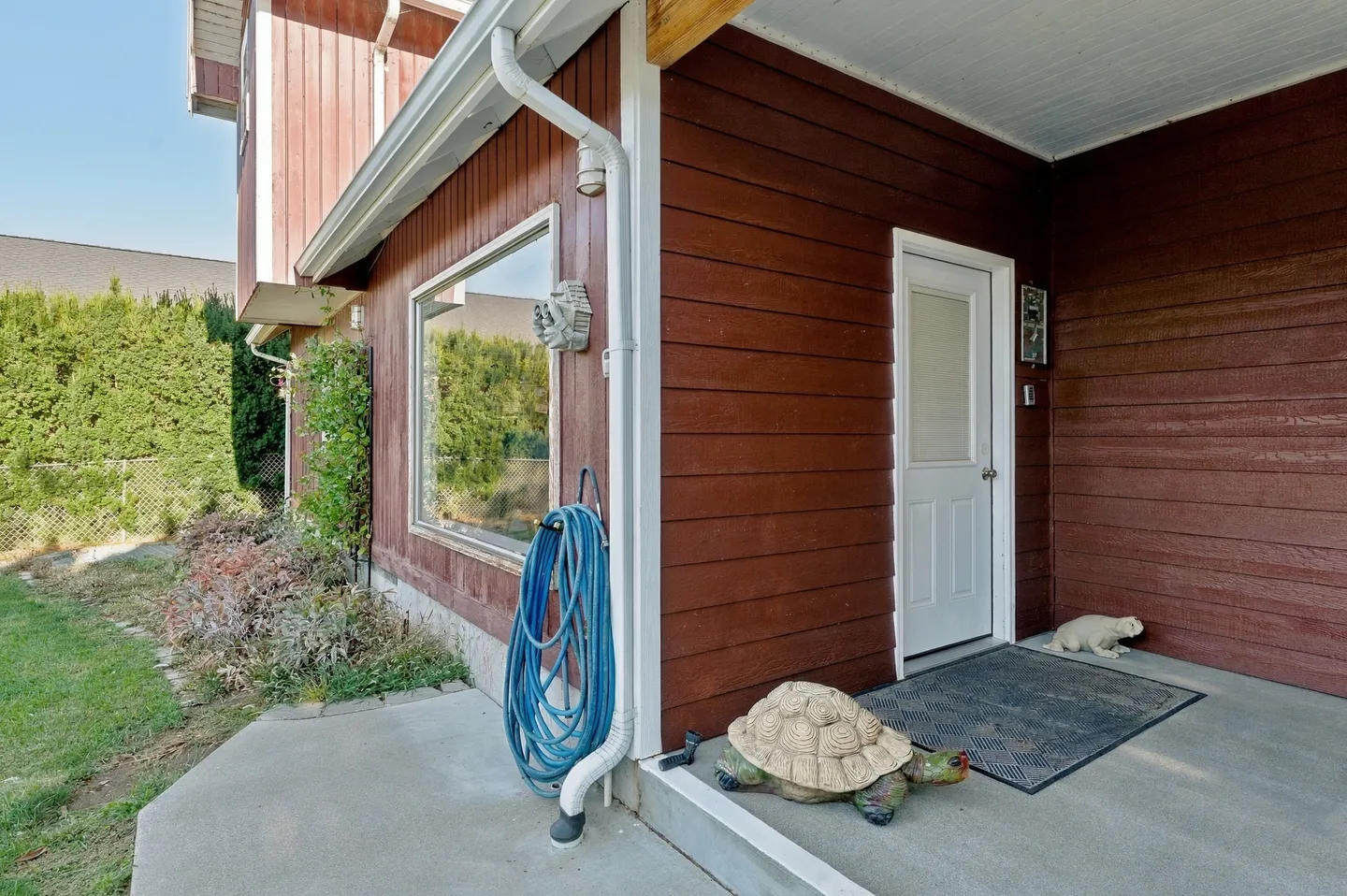 Exterior entryway of a home featuring rust-red horizontal siding, a white door, concrete porch, and a garden hose.