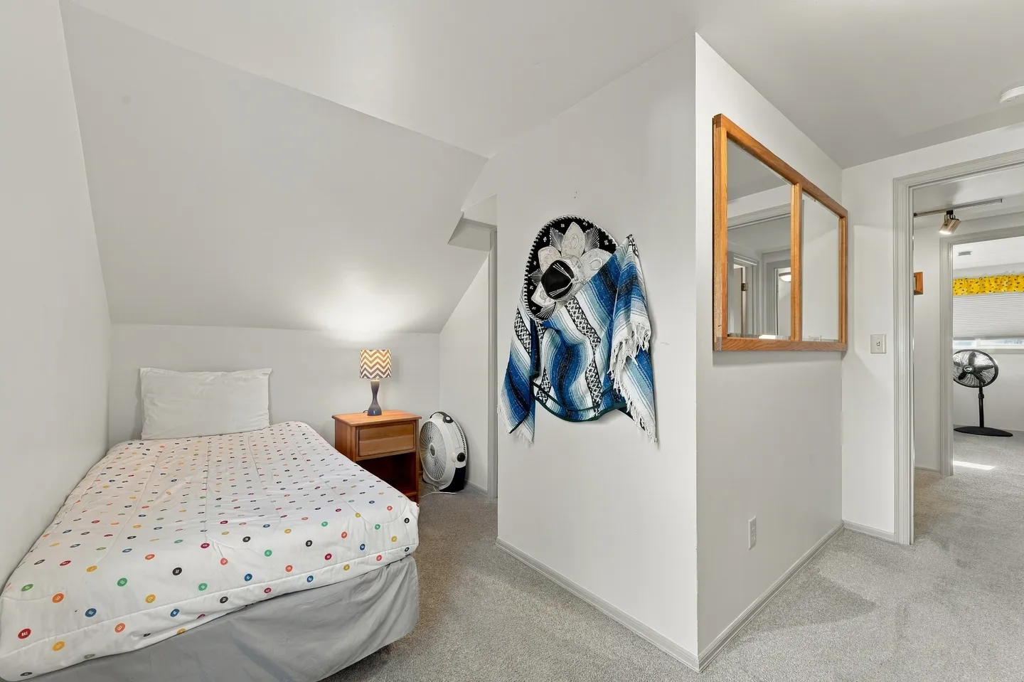 A small alcove bedroom with a single bed under a sloped ceiling, beside an interior mirror and hallway.