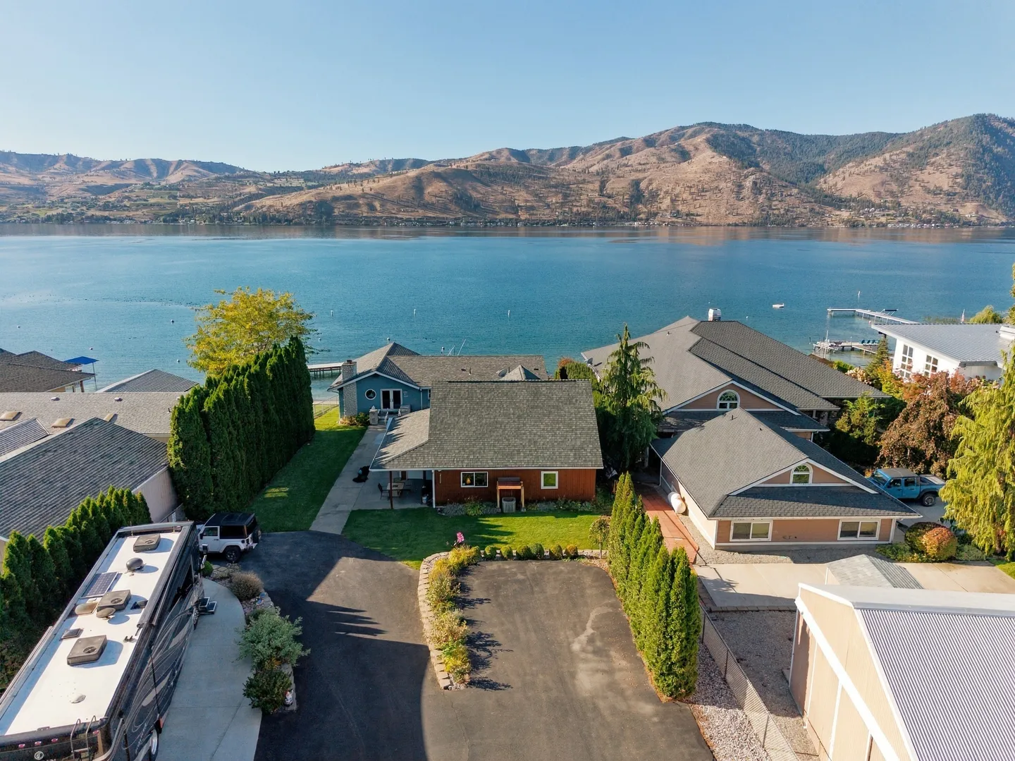Aerial view of a waterfront home with a paved driveway, front lawn, and views of a large lake and mountains.