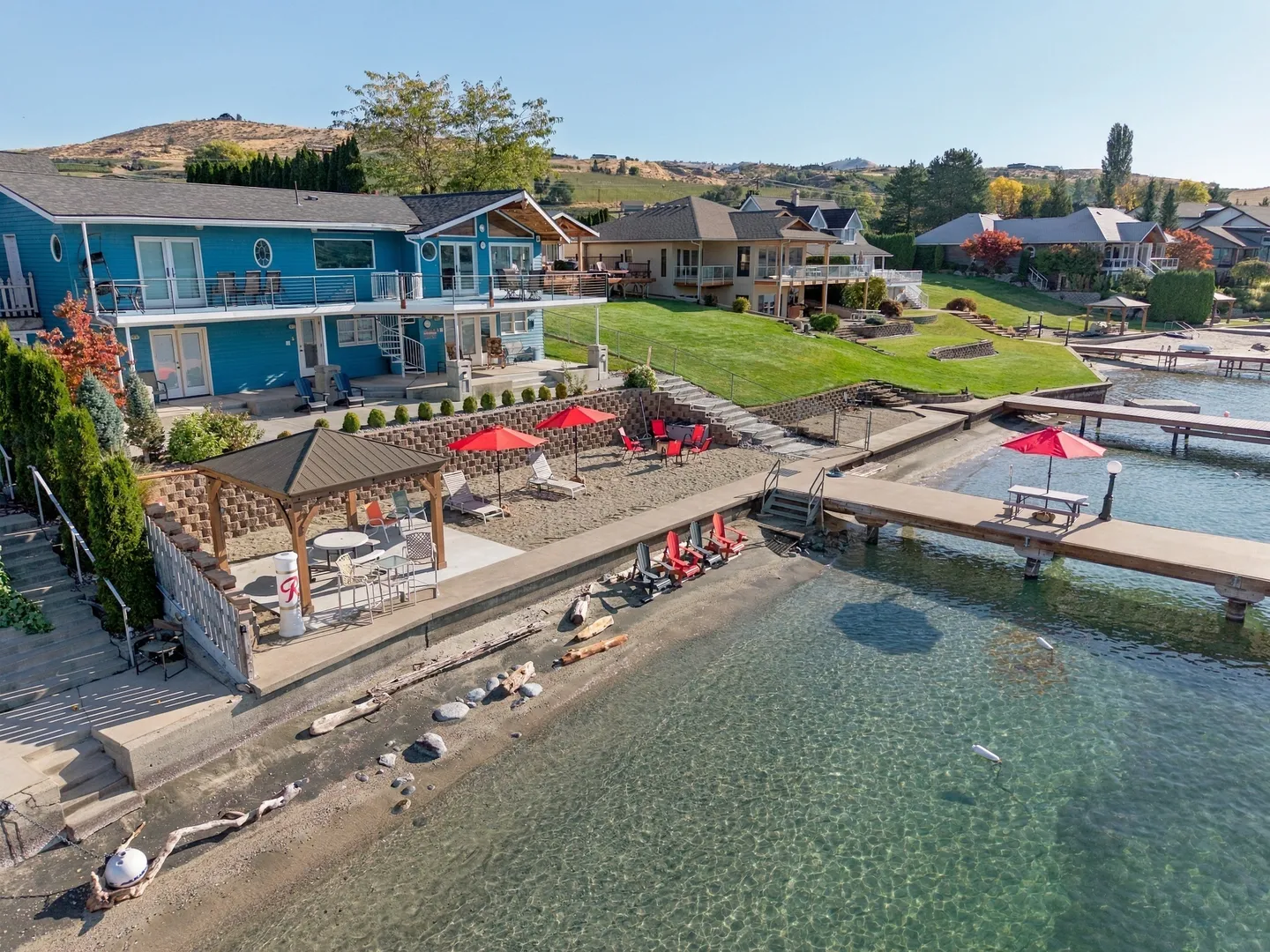 Aerial view of a waterfront home with a blue exterior, multilevel patio, gazebo, and private dock extending into clear water.