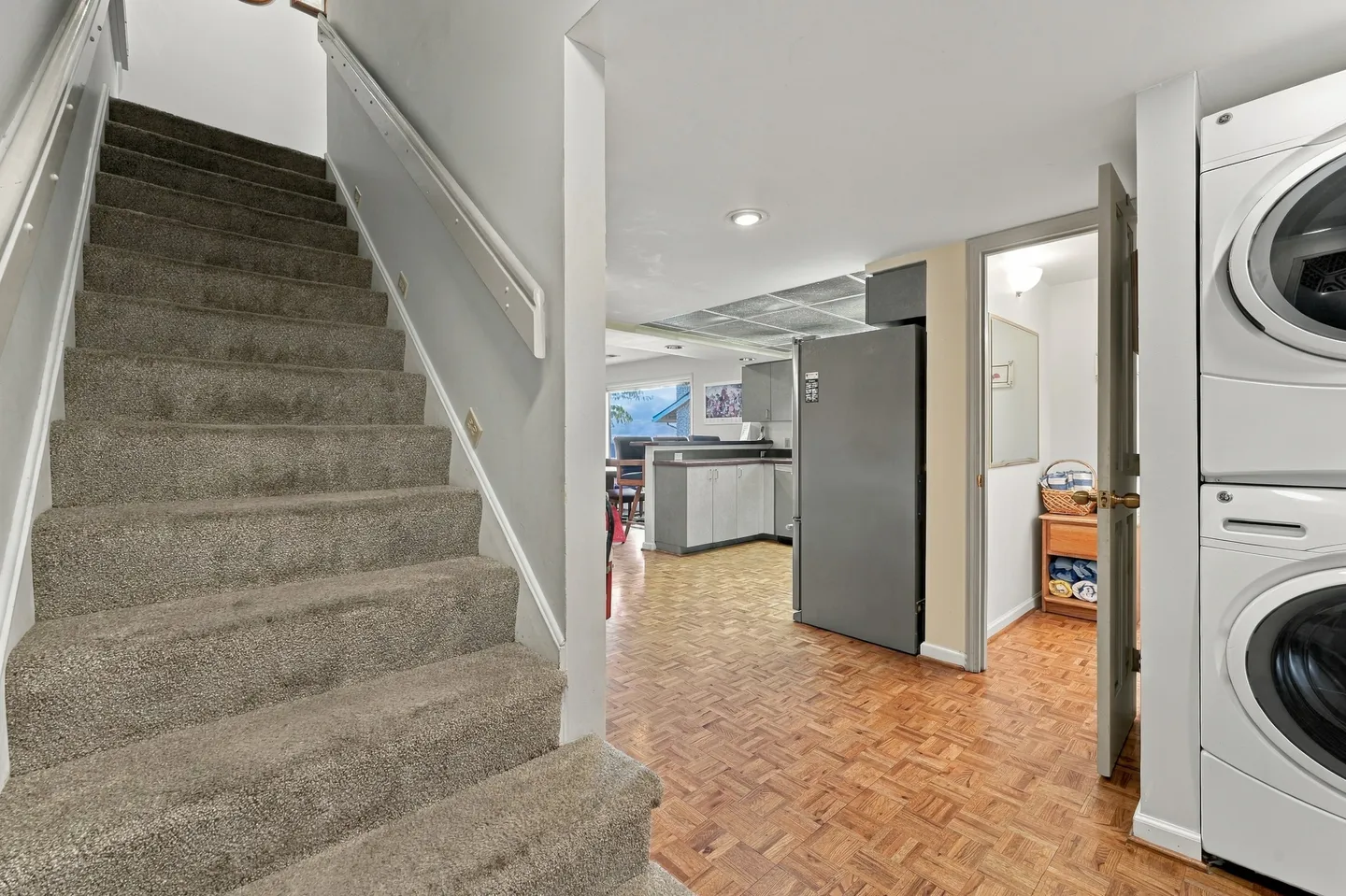 A carpeted staircase next to parquet flooring leading toward a kitchen, adjacent to a stacked washer and dryer.