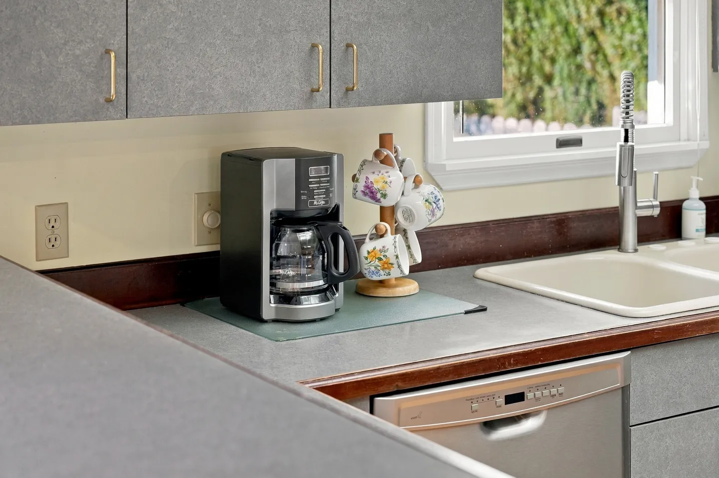 Close-up of a kitchen countertop showing a drip coffee maker, mug tree with floral cups, sink with tall faucet, and gray cabinets.