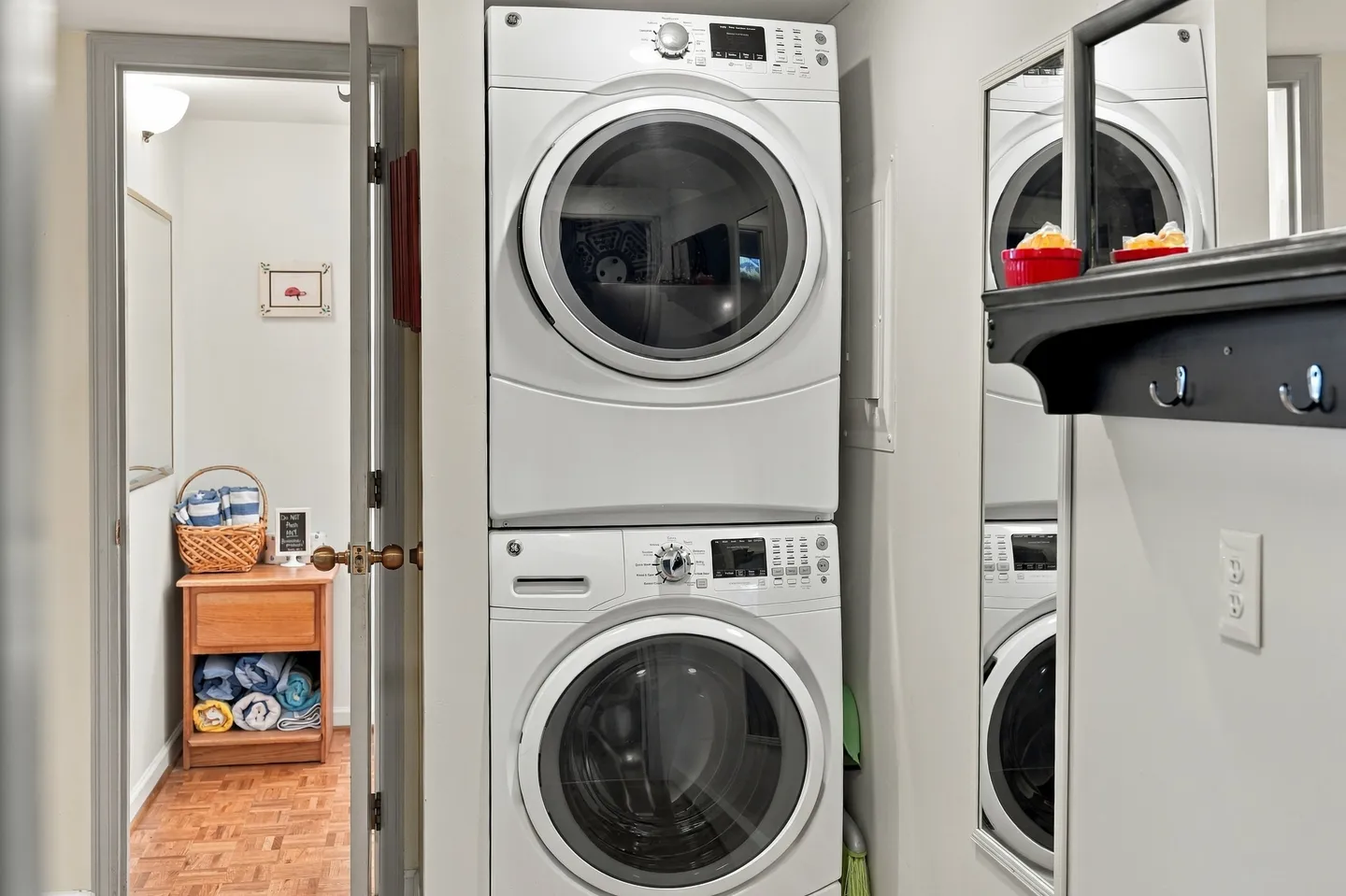 A stacked white washing machine and dryer set in an alcove near a hallway doorway and mirror.