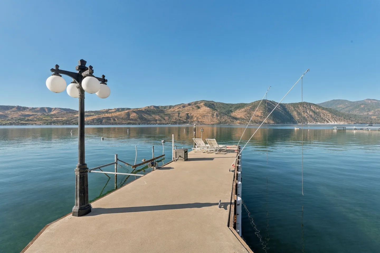 A long concrete dock extending into a wide lake with lounge chairs at the end, set against mountain scenery under a clear blue sky.
