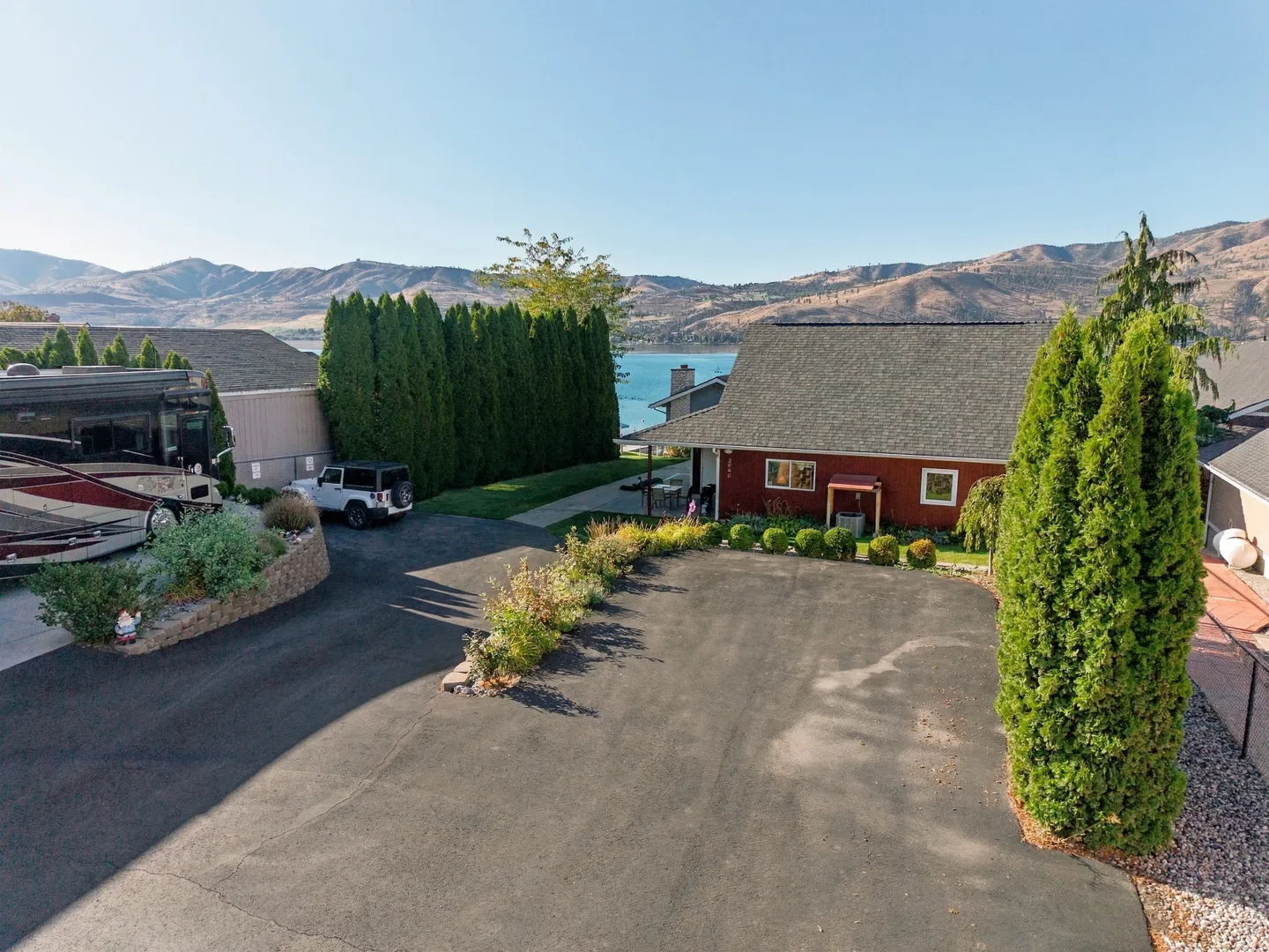 Elevated view of a wide paved driveway leading to a red house and an RV parked nearby, with a lake and mountains in the background under clear skies.