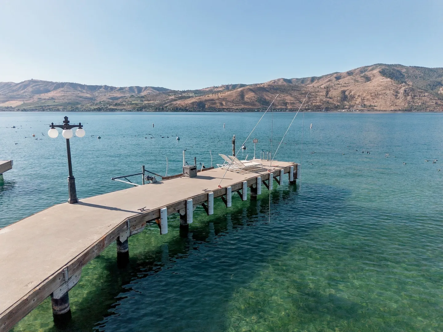 A long dock extending into clear turquoise water against a backdrop of hills across a lake.