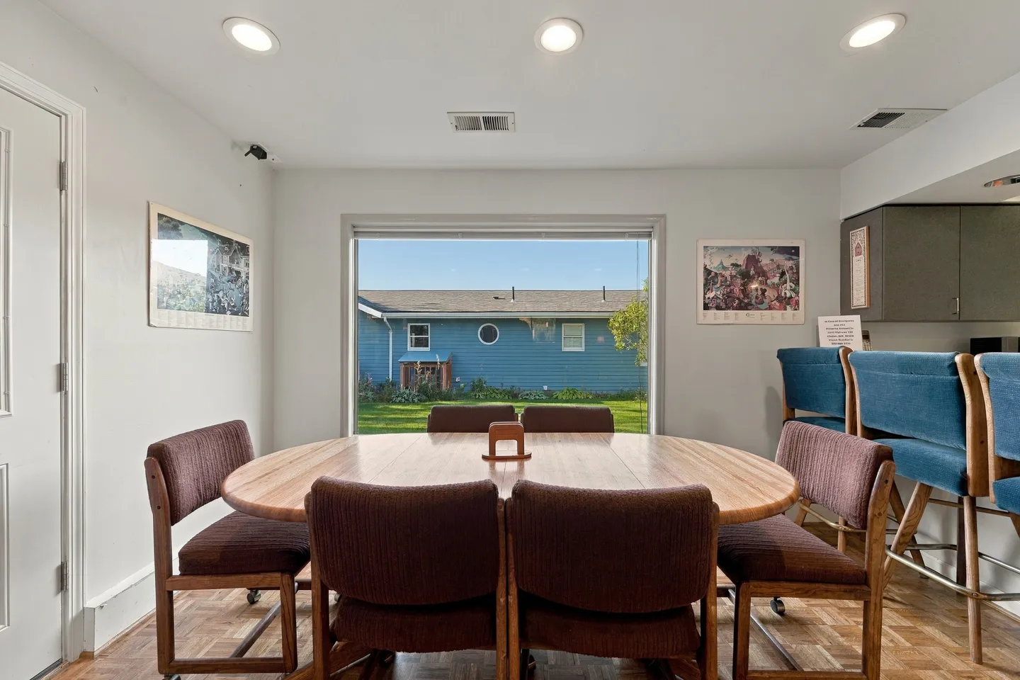 Dining area featuring an oval wooden table surrounded by upholstered chairs in front of a picture window overlooking a yard.