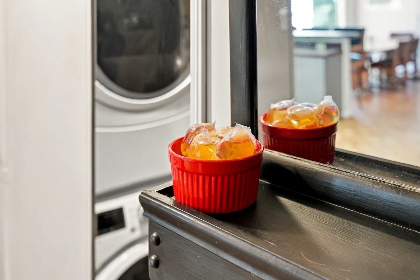 A red ceramic ramekin filled with laundry detergent pods sitting on a dark wooden ledge in front of a mirror with stacked laundry machines in the background.