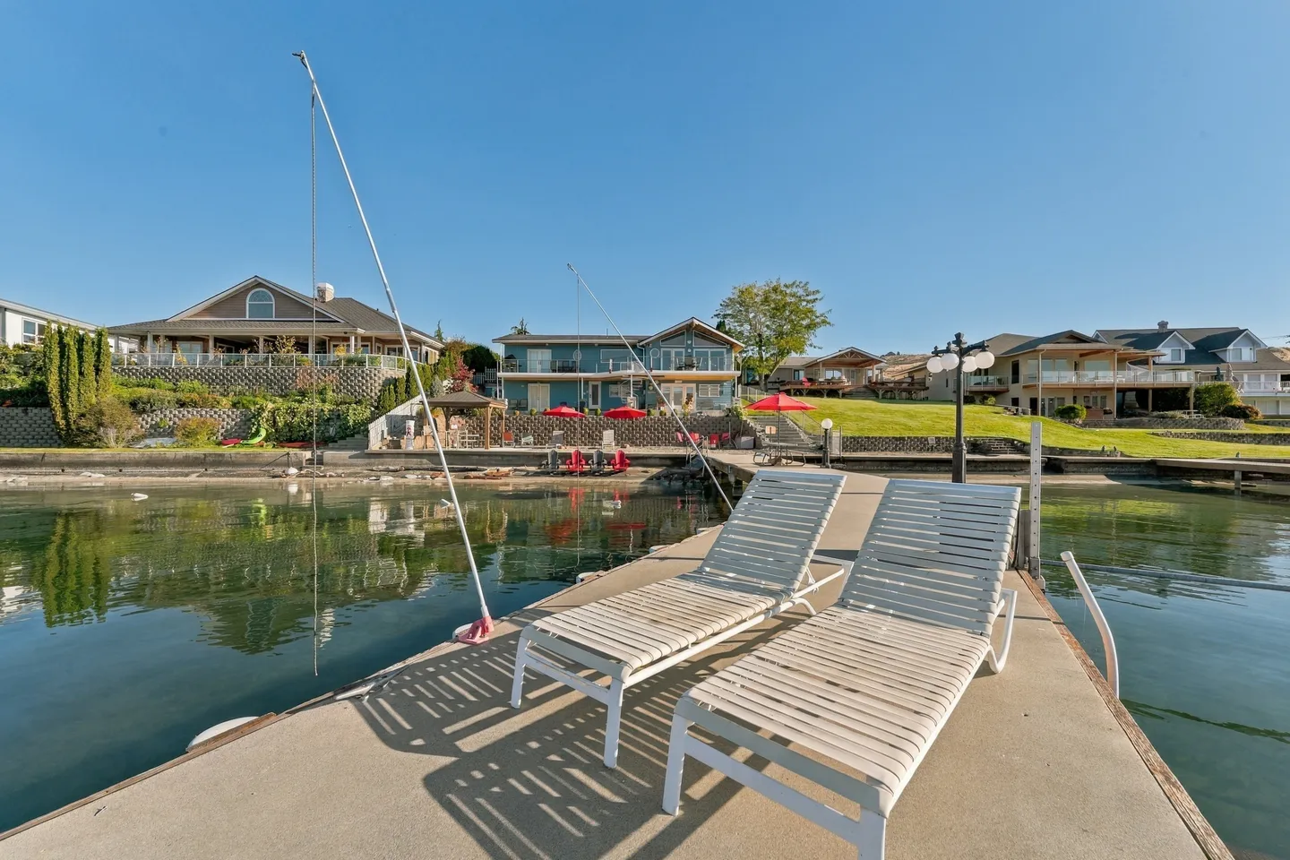 A floating concrete dock with two lounge chairs overlooking lakeside homes under a clear blue sky.