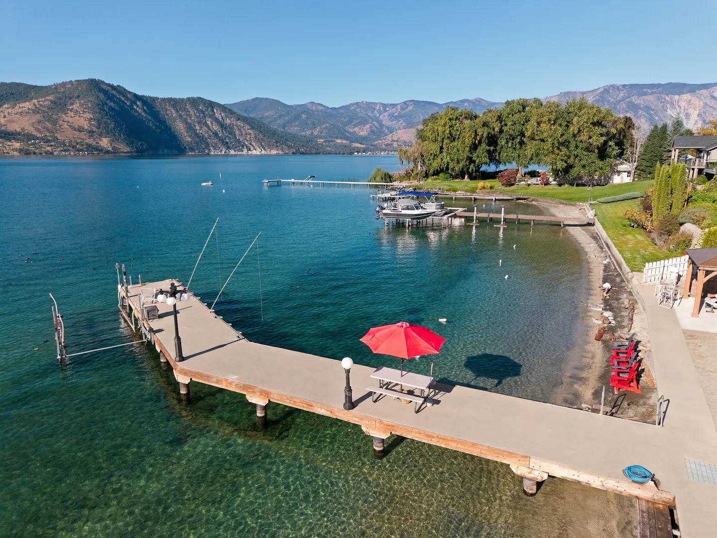Aerial view of a long concrete pier extending into clear lake water with mountains in the background.