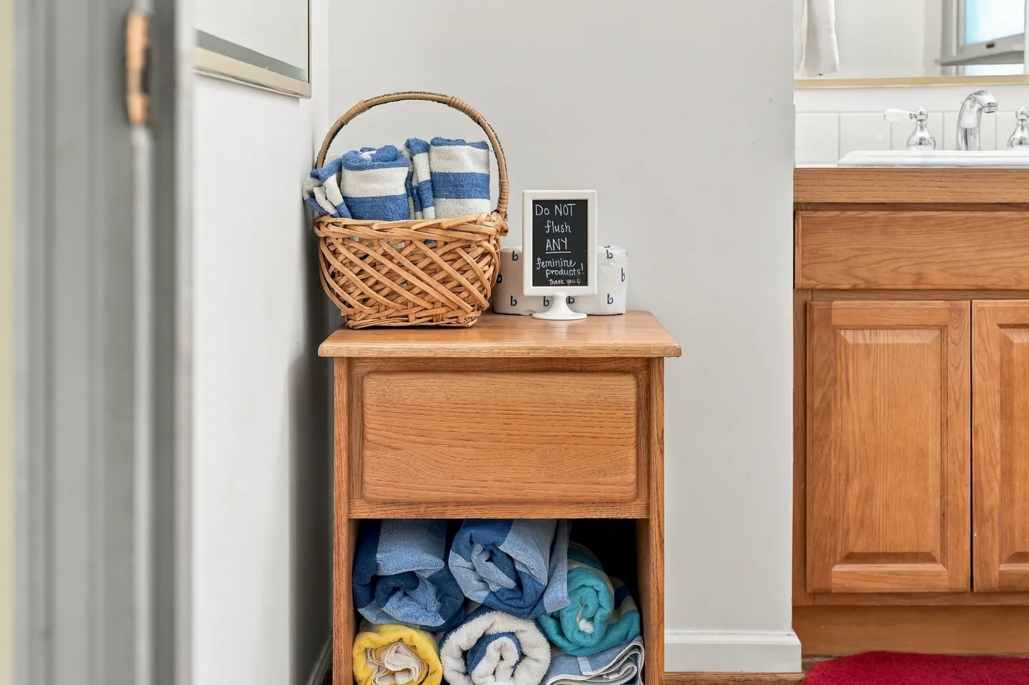 A wooden nightstand in a bathroom holding extra rolled towels, a wicker basket with towels, and rolls of toilet paper next to a vanity.
