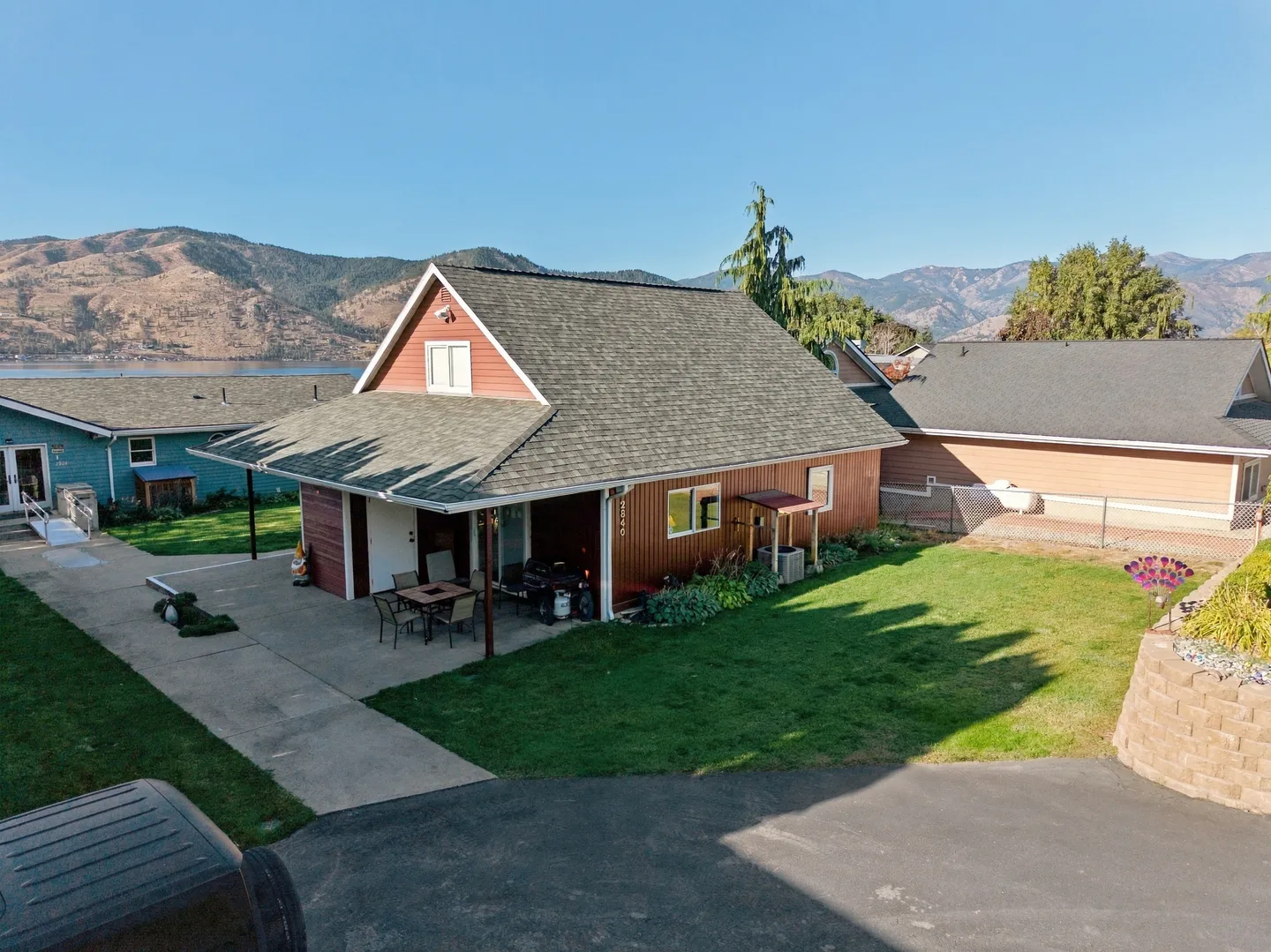 Aerial view of a red-sided home with a grassy lawn, driveway, covered patio, and mountains in the background behind a body of water.