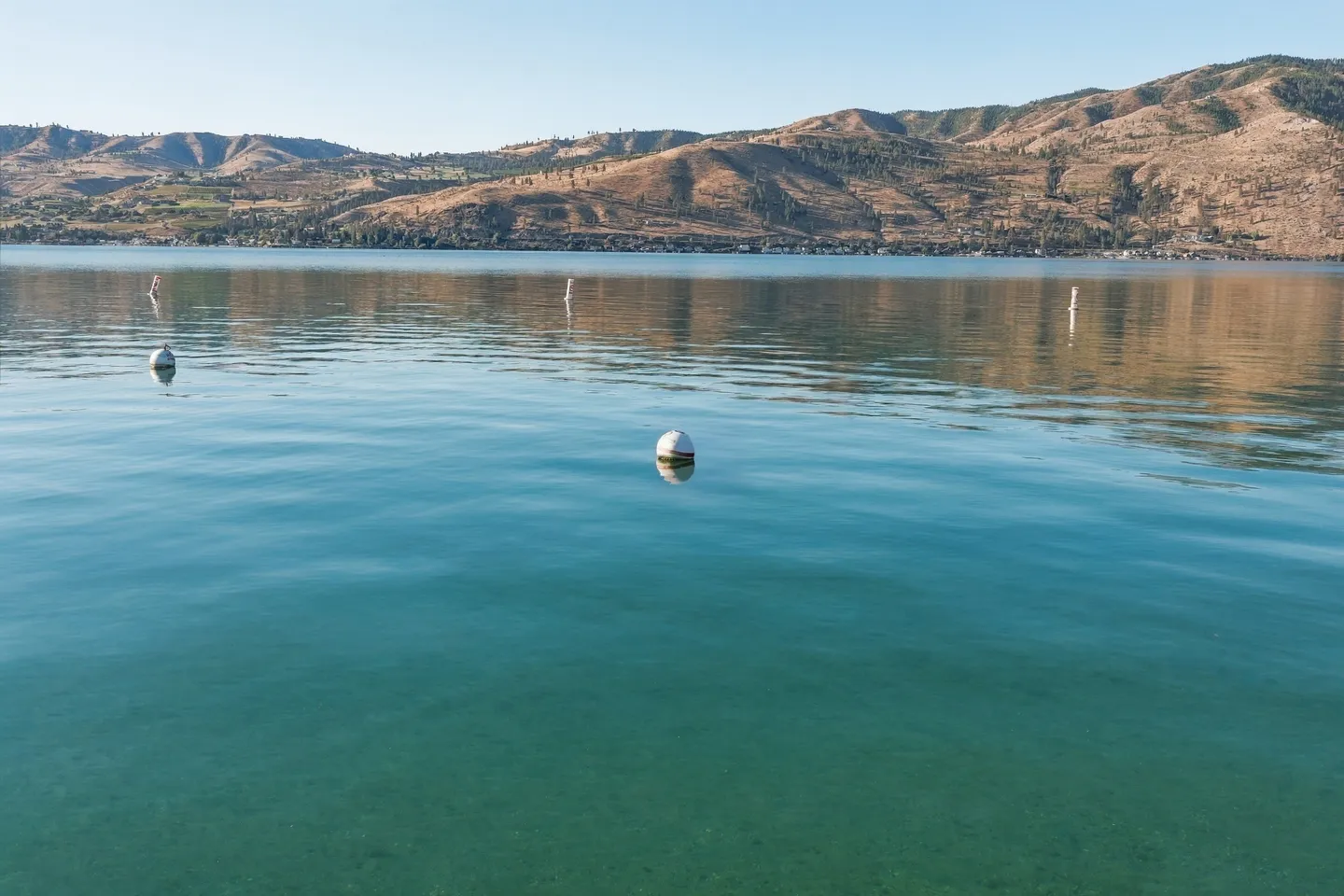 Calm, clear blue water of a lake with buoys floating near the surface and arid hills in the background.