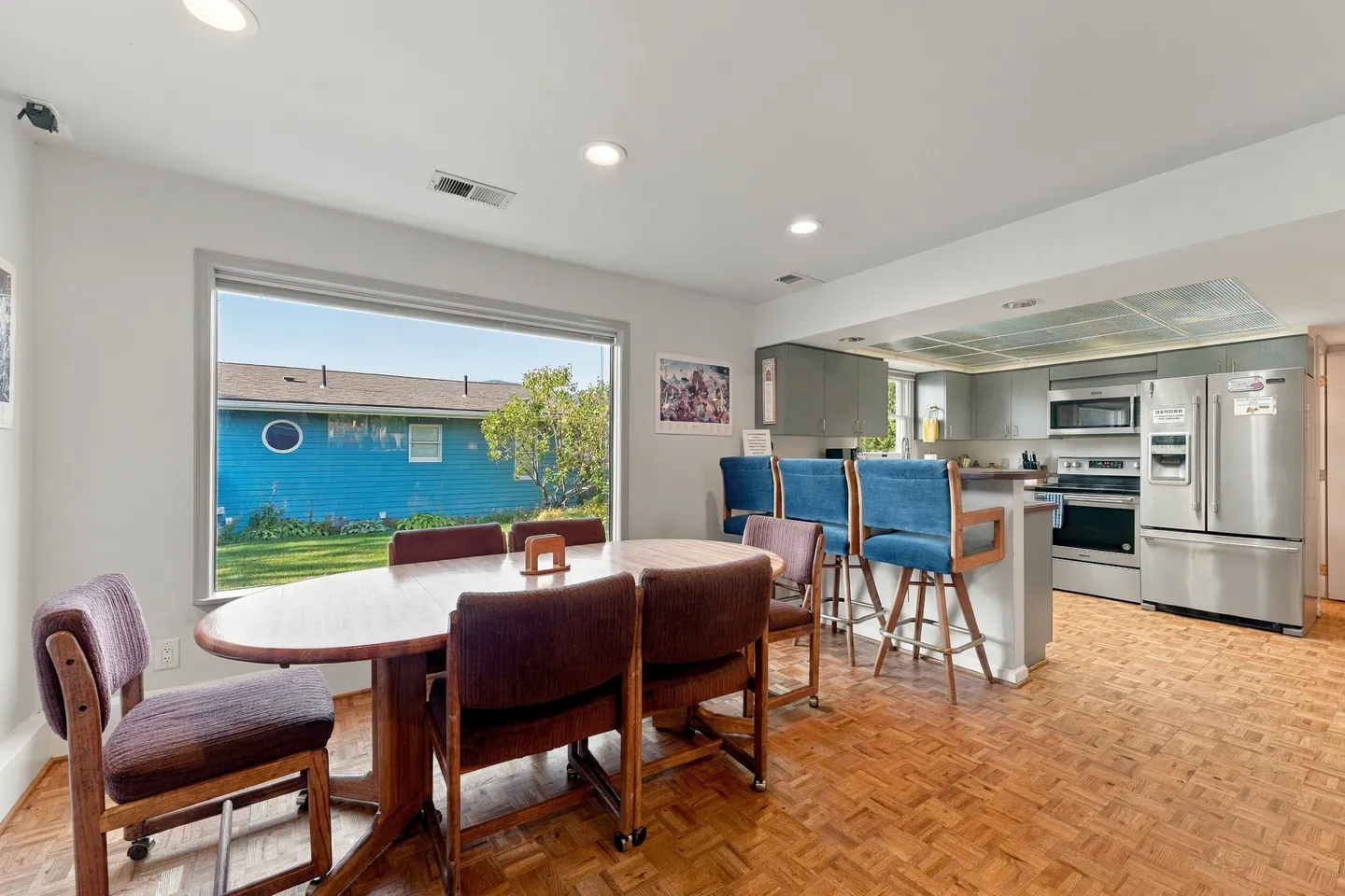 Dining area with an oval table and rolling upholstered chairs next to a large picture window, adjacent to an open kitchen with bar seating.