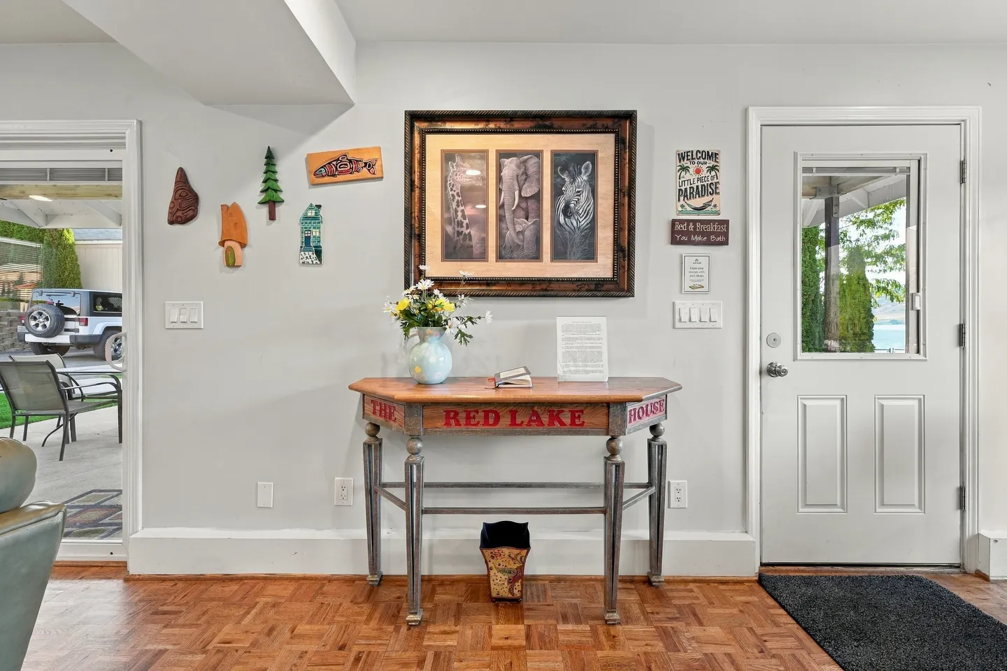 An entryway featuring parquet flooring, an exterior door with a window, and a wooden console table labeled "The Red Lake House" beneath wall art.