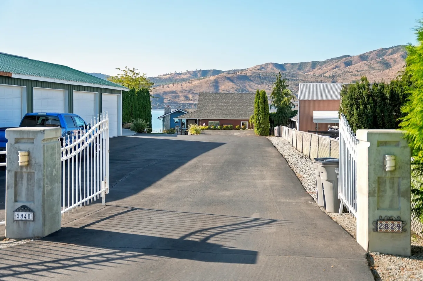 Open white iron gates leading into a wide paved driveway toward a home with lake and mountain views in the background.