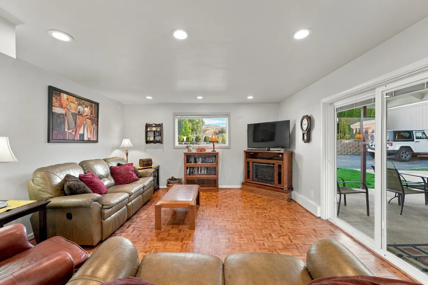 Living room with leather seating, wooden flooring, a corner television stand with fireplace insert, and sliding glass doors to the outside.