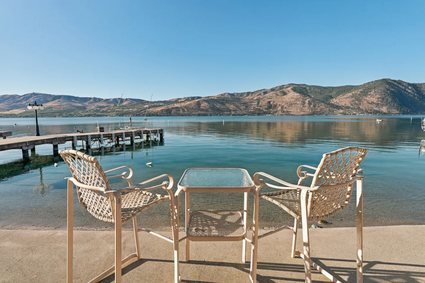Two outdoor armchairs and a small glass-topped table set on a concrete patio overlooking a calm lake and distant hills with a wooden dock nearby.
