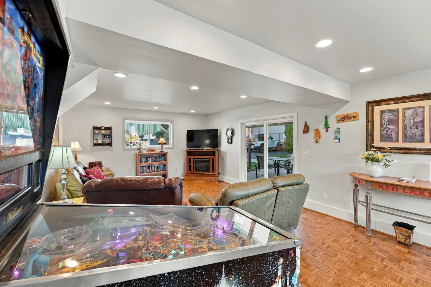 Game room and lounge area with a pinball machine in the foreground, leather sofas, a TV on a console, and sliding glass doors leading outside.