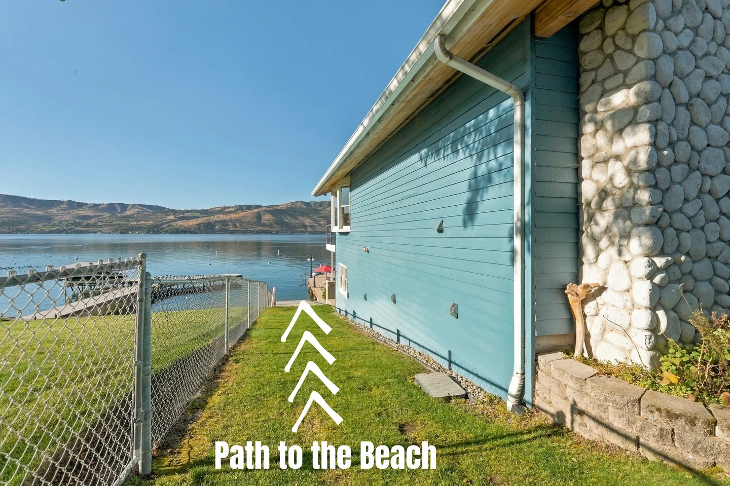 A grassy side yard path next to a light blue house leading down to a lake and dock.