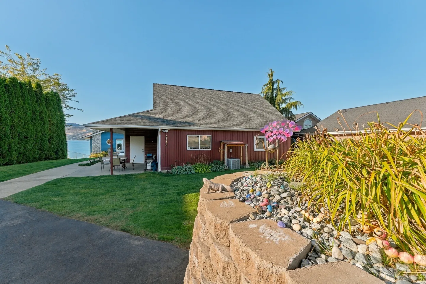 Exterior view of a red siding home with a paved driveway, curved stone retaining wall, and water visible in the background.