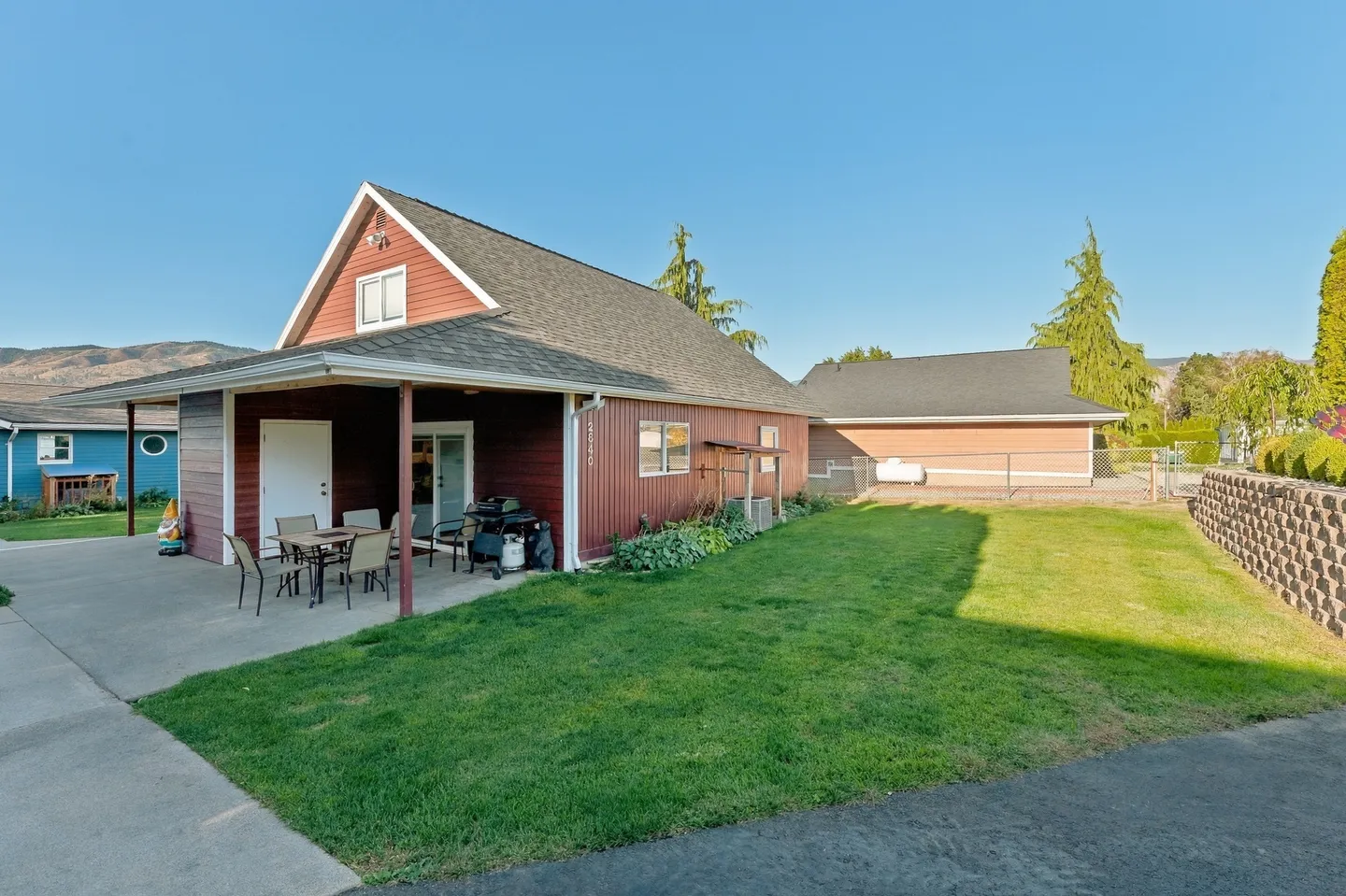 Exterior backyard view of a red-sided home with a covered concrete patio, lawn, and surrounding residential neighborhood.