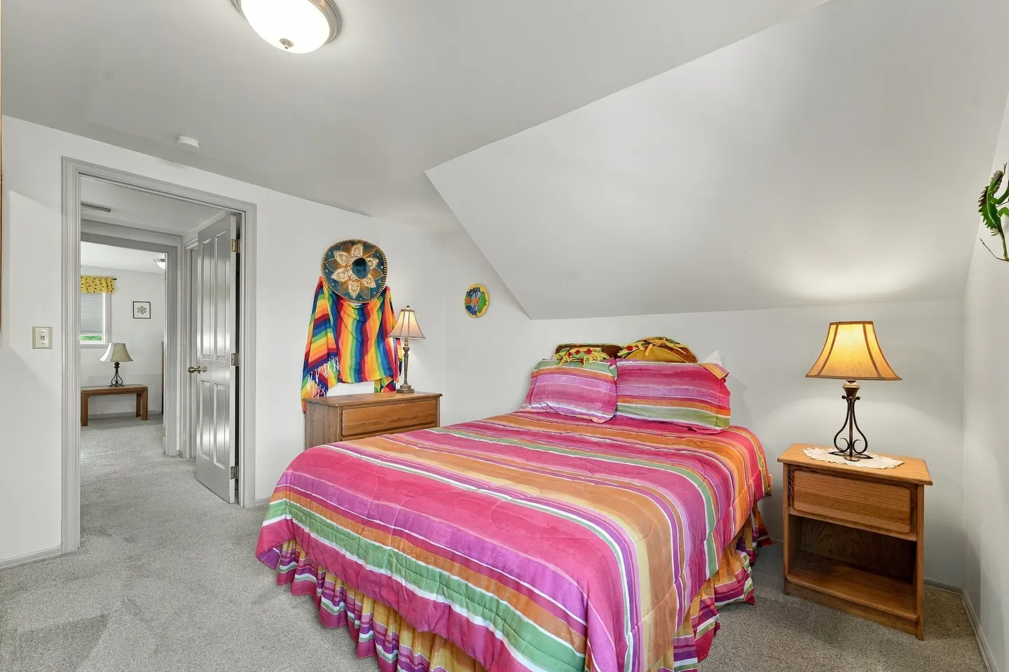 Bedroom with a sloped ceiling, carpeted floor, bed with pink striped bedding, nightstands, and a doorway to a hallway.