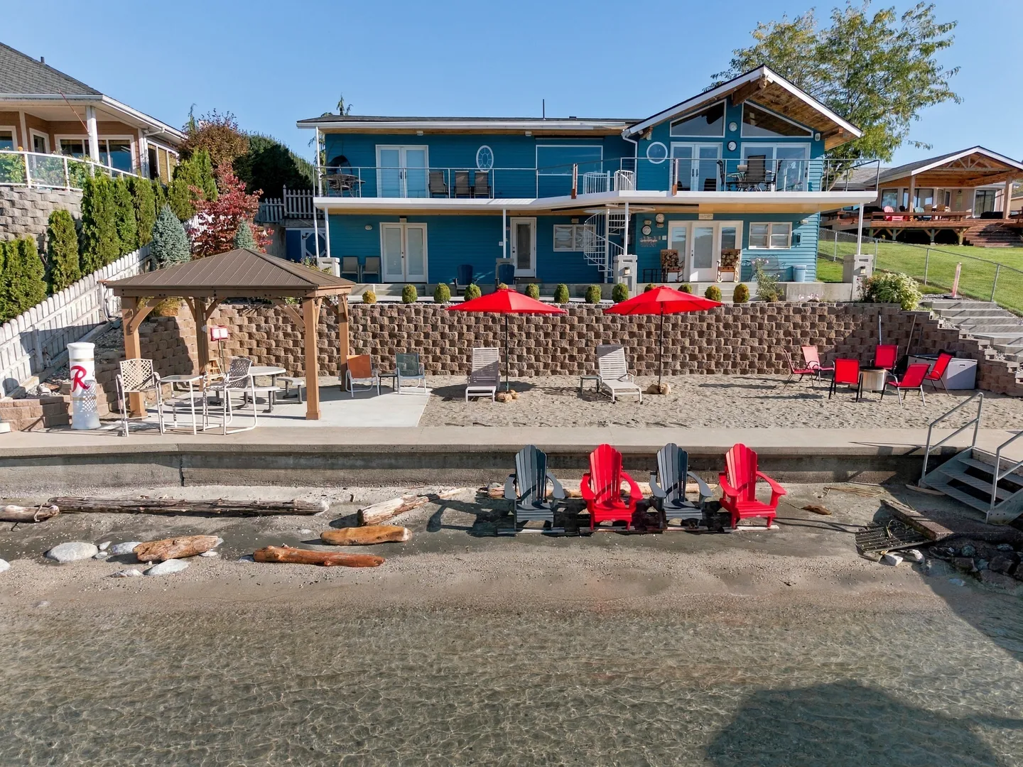 Exterior view of a two-story blue lakefront home with a tiered sandy beach, patio, gazebo, and chairs by clear water.