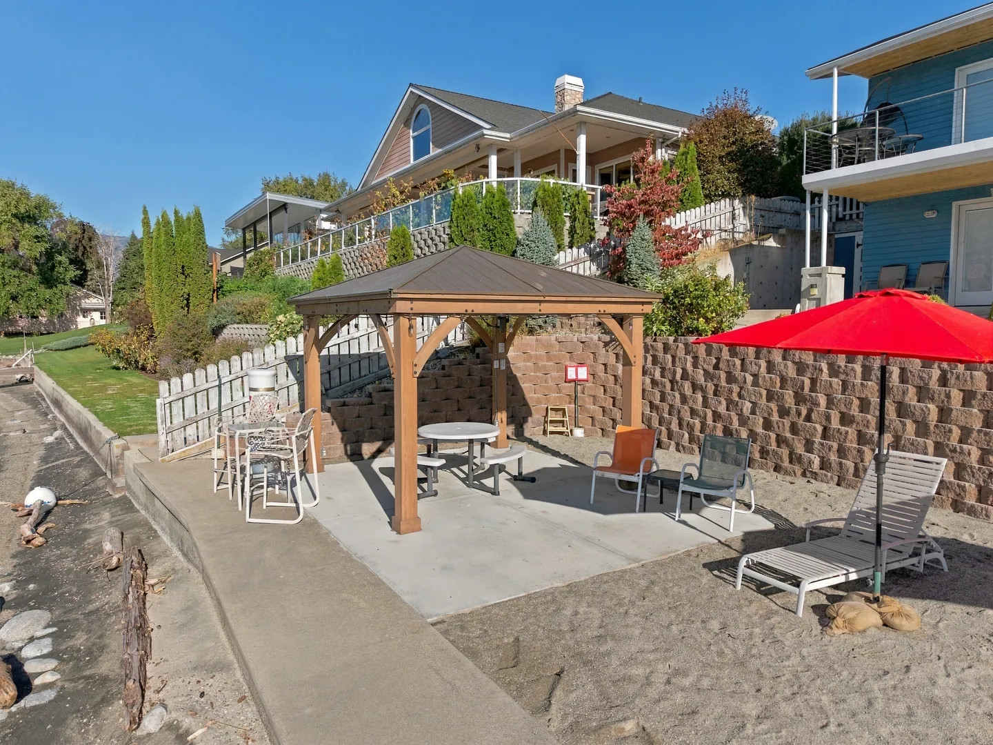 Outdoor beach patio featuring a wooden gazebo with a picnic table, patio chairs, a red umbrella, and a tiered retaining wall leading up to a hillside home.
