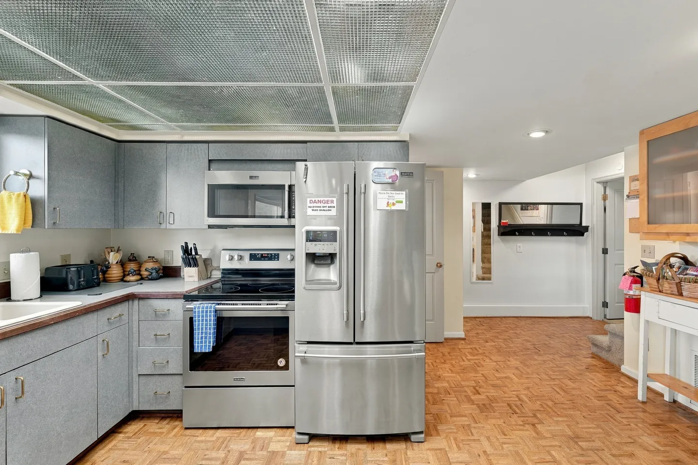 Kitchen with stainless steel refrigerator, microwave, and stove alongside parquet-style flooring and textured ceiling panels.