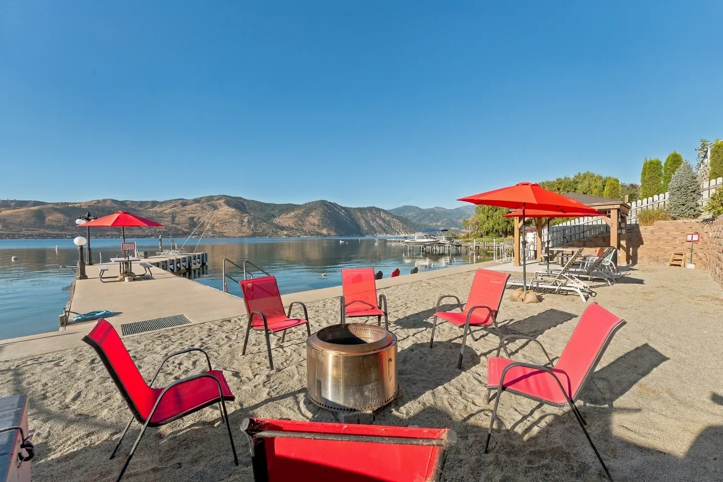 A sandy beachfront seating area with red chairs arranged around a circular stainless steel fire pit, overlooking a calm lake and mountains.