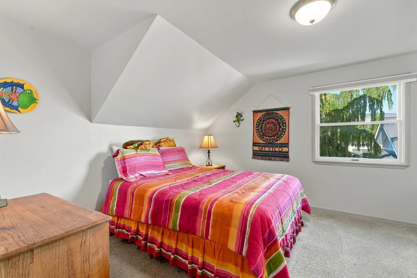 Bedroom with an angled ceiling, a bed made with colorful striped bedding, carpeted floor, and a window.