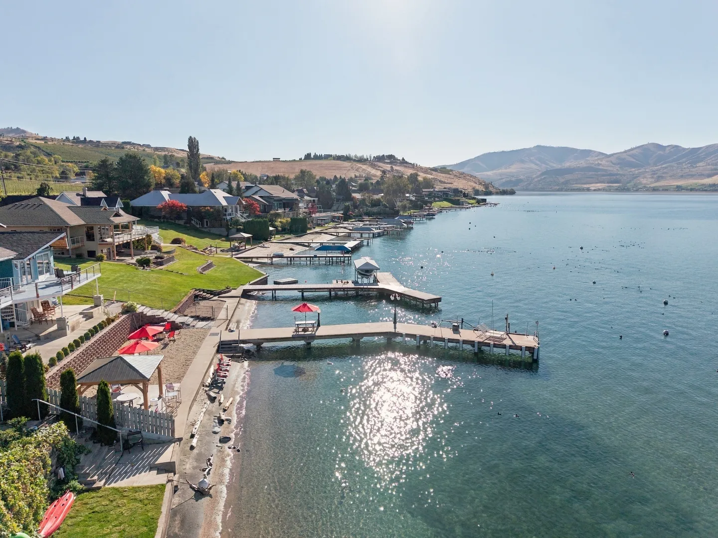 Aerial view of a lakefront shoreline with private wooden docks, outdoor patios, grassy lawns, and houses beneath a clear sunny sky.